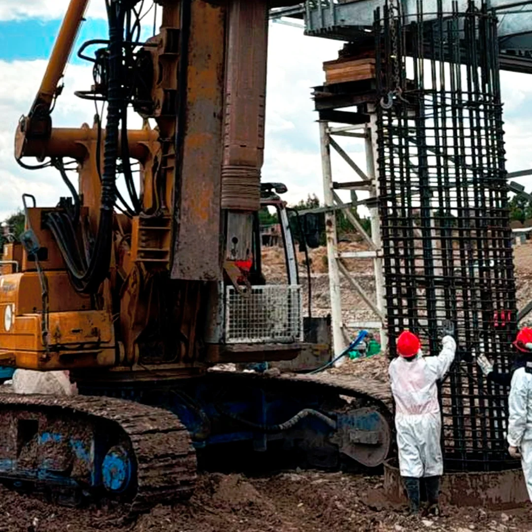 Colocación de armadura de acero cilíndrica en pozo de pilote excavado, con perforadora rotativa y dos operarios de DCC Perú durante ejecución de cimentación profunda de puente