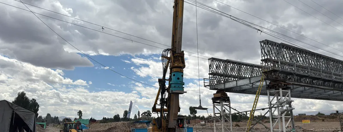 Equipo de perforación ejecutando los pilotes excavados del Puente Cunas, con la estructura del puente al fondo