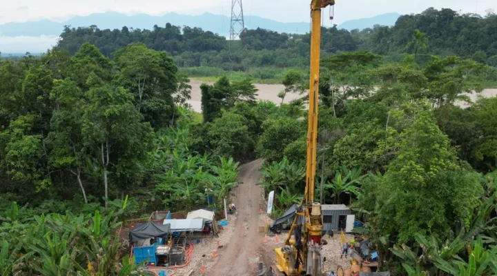 Equipo de perforación con el mástil vertical en una plataforma abierta en la selva, con la torre de alta tensión al fondo