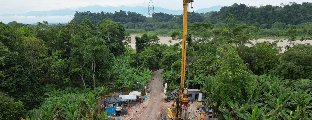 Equipo de perforación con el mástil vertical en una plataforma abierta en la selva, con la torre de alta tensión al fondo